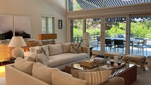 Un salón amplio con un sofá grande, una mesa de centro y un ventilador de techo. Hay un ventanal con persianas y vistas al exterior.