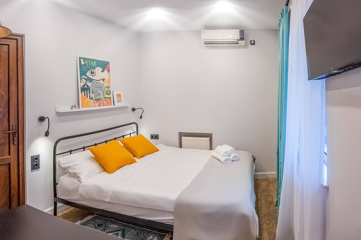 A neatly made bed with white and orange bedding, a wall-mounted air conditioner, and a framed picture above the bed.