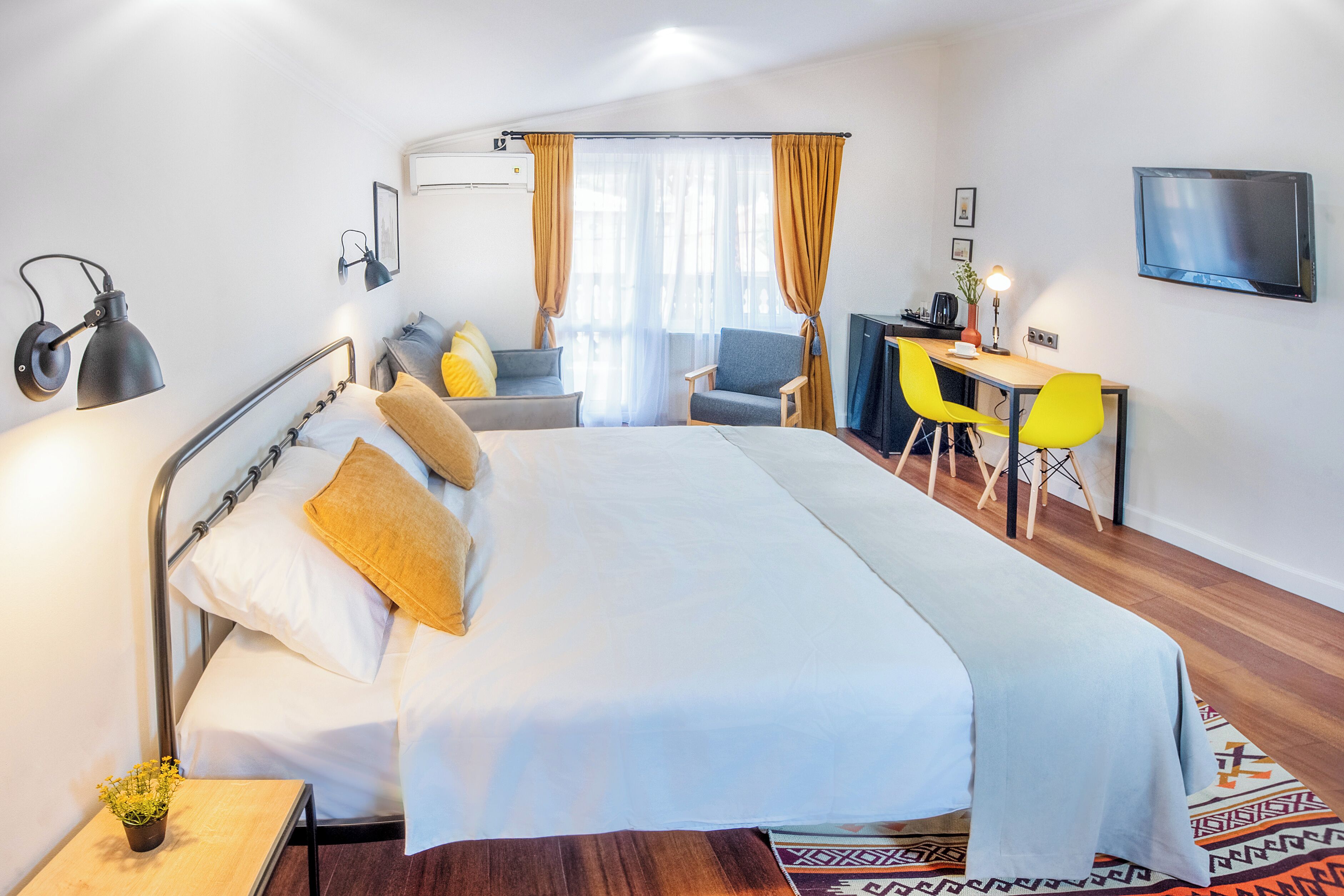 A hotel room with a bed, desk, two chairs, a TV, and a small table with a plant.