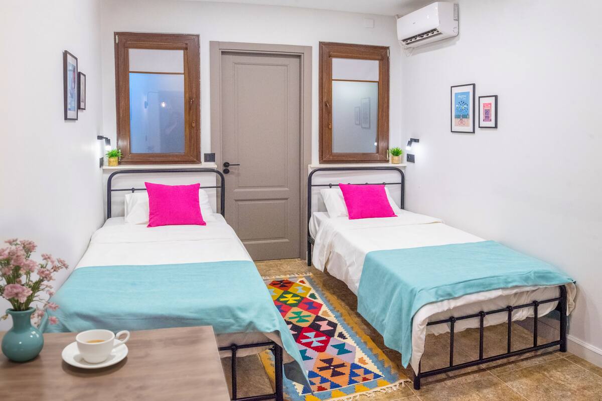 A room with two single beds, a small table with a cup, and a colorful rug.