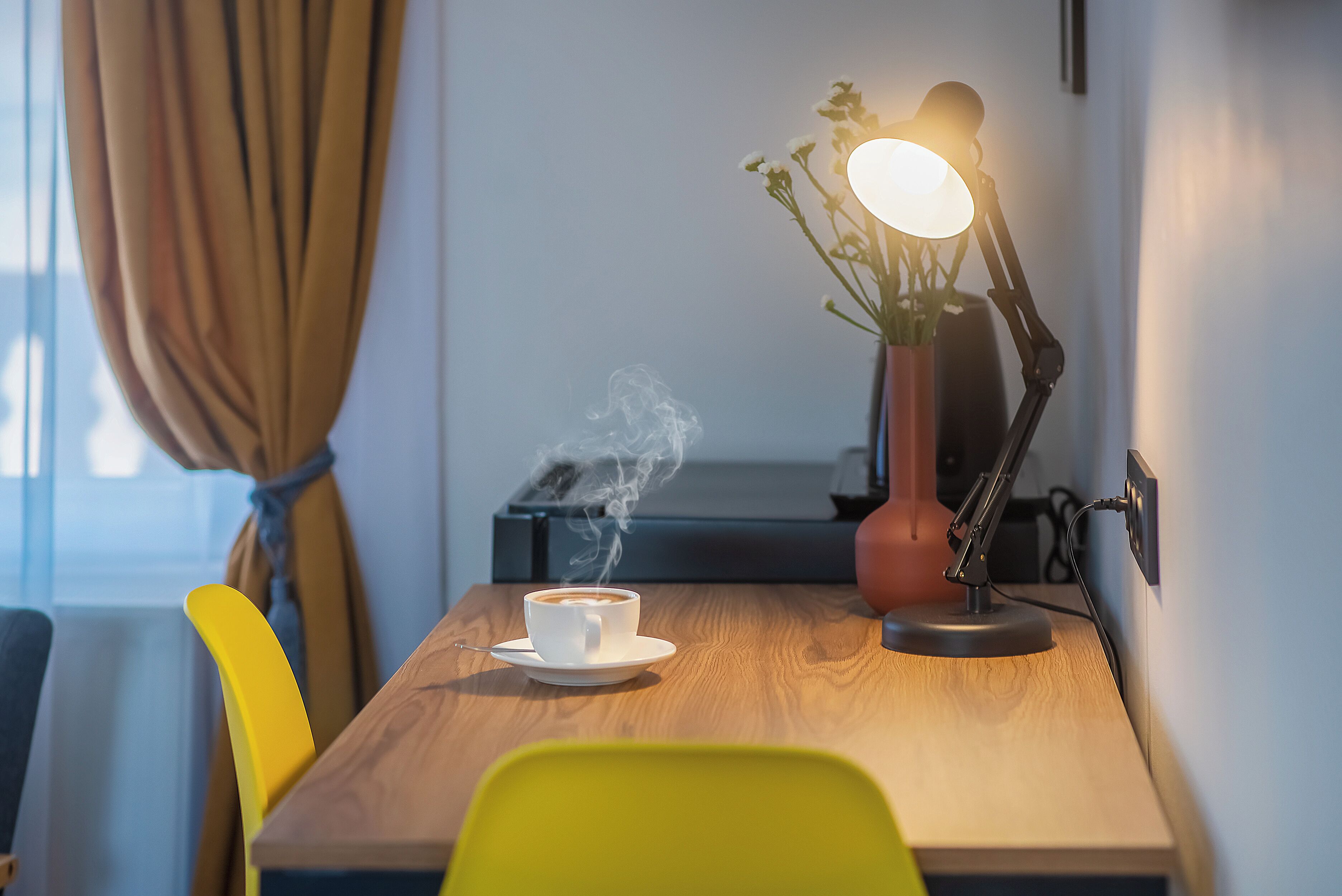 A wooden table with a white cup of coffee emitting steam, a yellow chair, a desk lamp, and a potted plant.
