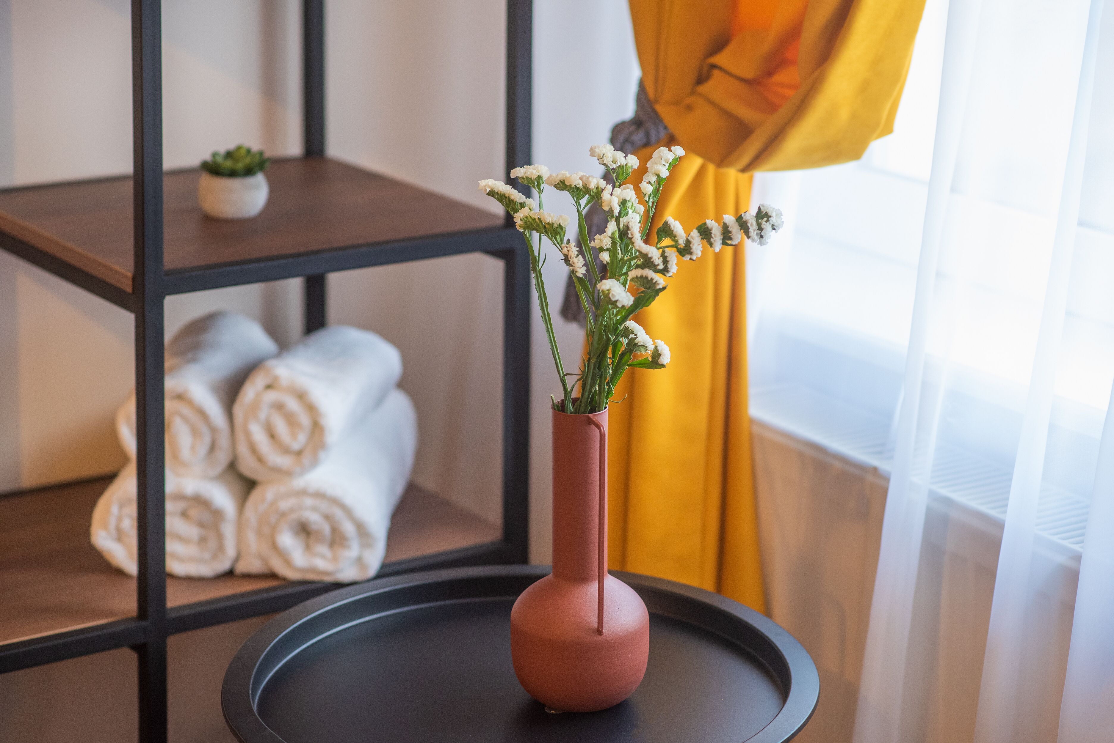 A vase with flowers on a table, a shelf with rolled towels, and a window with curtains.