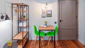 Una habitación pequeña con una mesa de madera, dos sillas verdes, un estante metálico, una puerta y cuadros colgados en la pared.
