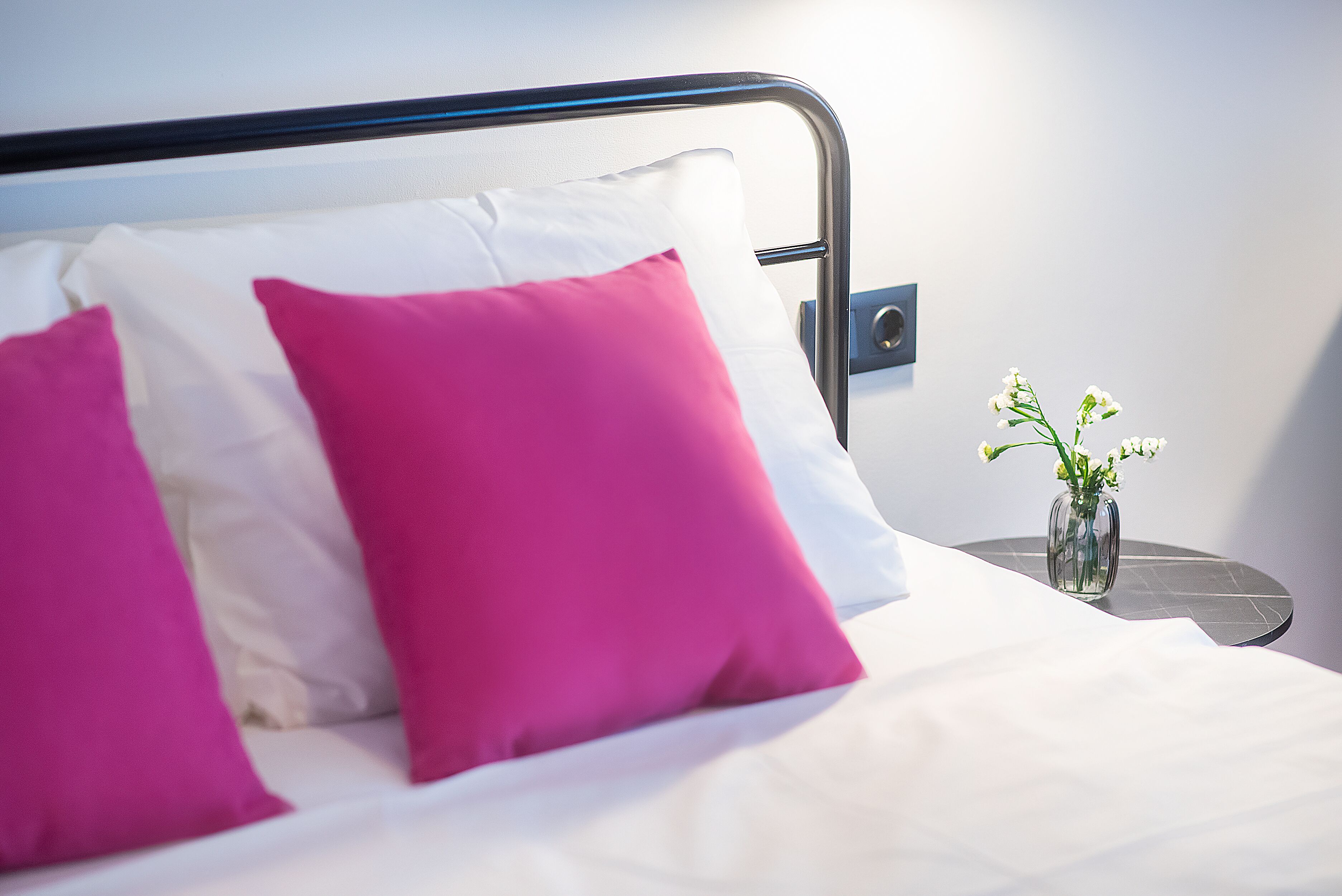 Un lit avec des oreillers blancs et roses, une petite table ronde sur laquelle est posé un vase de fleurs, et un interrupteur fixé au mur.