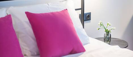 Un lit avec des oreillers blancs et roses, une petite table ronde sur laquelle est posé un vase de fleurs, et un interrupteur fixé au mur.