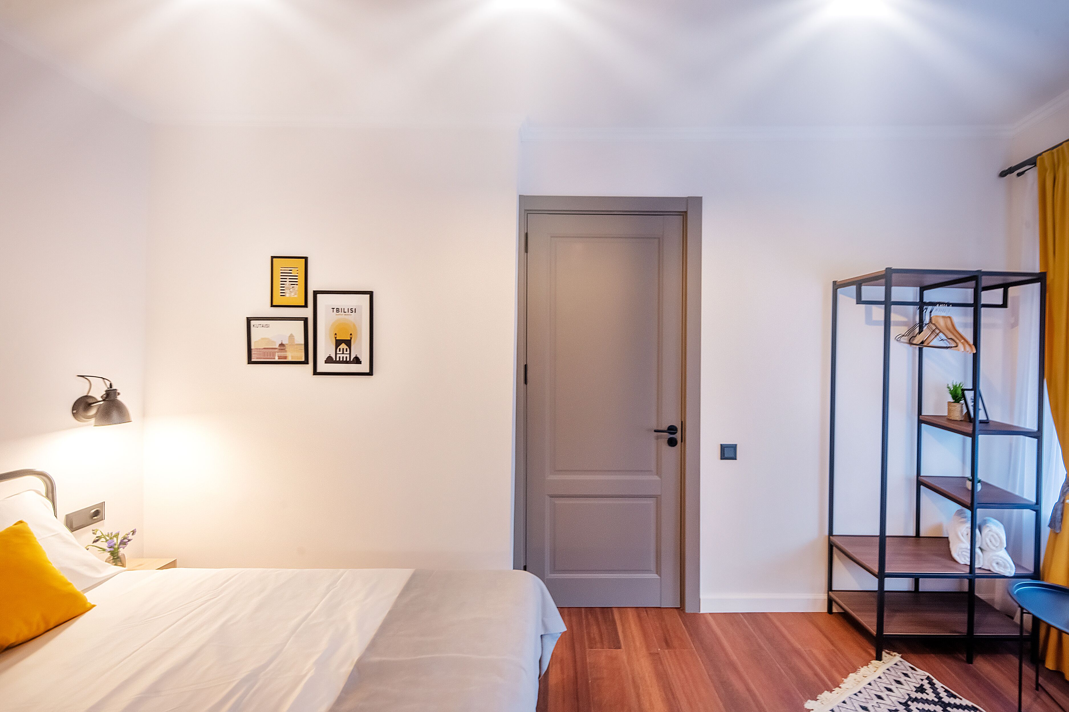 A bedroom with a bed, a nightstand, a wall-mounted lamp, a door, and a shelf with towels and a plant.
