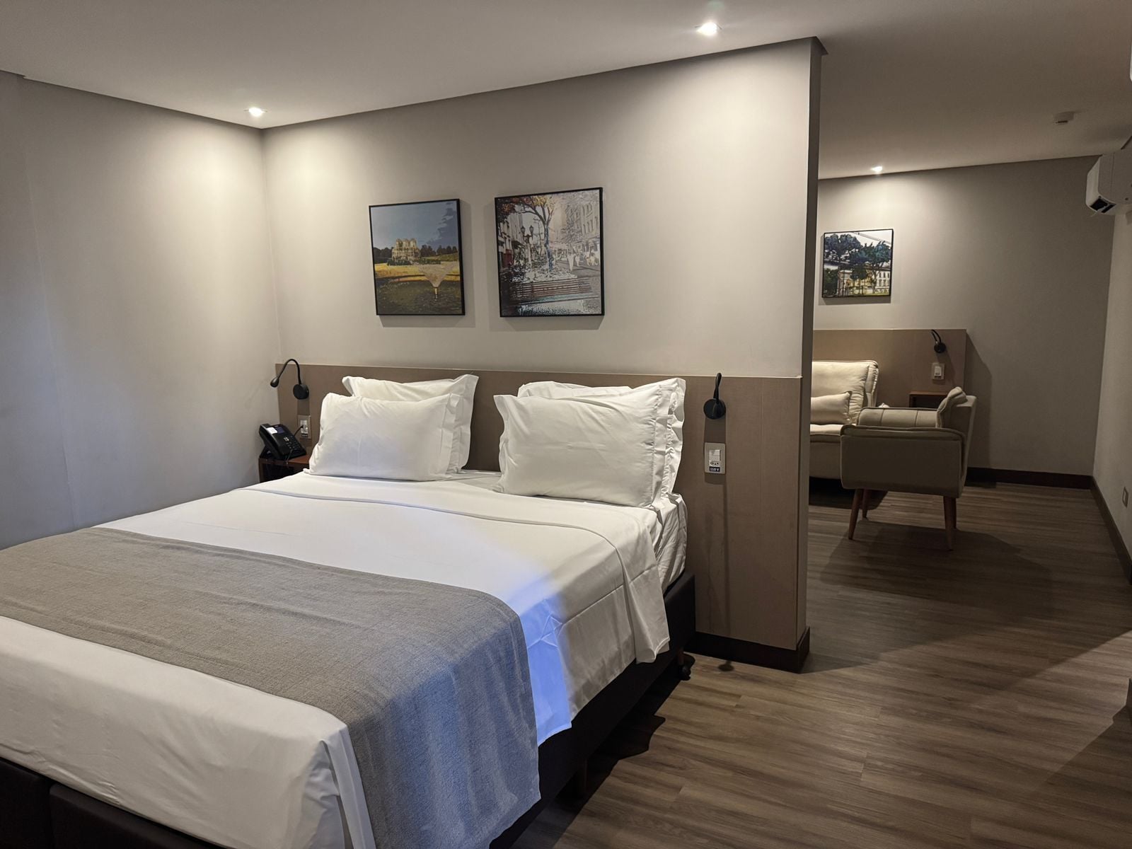 Ett modernt hotellrum med en stor säng, ett skrivbord och en stol. Det finns två tavlor på väggen och ett litet bord med en lampa.
