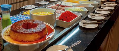 A buffet with various food items including a flan, fruits, and a green drink.