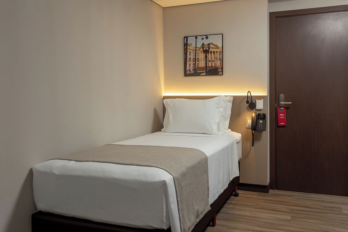 Une chambre d’hôtel avec un lit, un téléphone sur la table de chevet, une porte avec une carte rouge et un tableau accroché au mur.