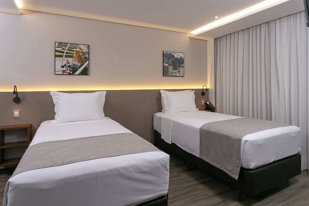 Une chambre d’hôtel avec deux lits simples, une table de chevet, une lampe fixée au mur et deux tableaux encadrés.