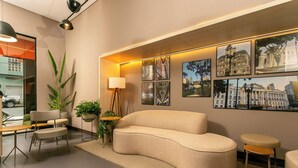 Un salon moderne avec un canapé aux lignes courbes, une table basse et une table d’appoint sur laquelle se trouve une lampe. Des tableaux encadrés ornent le mur, et des plantes en pot y sont également disposées.