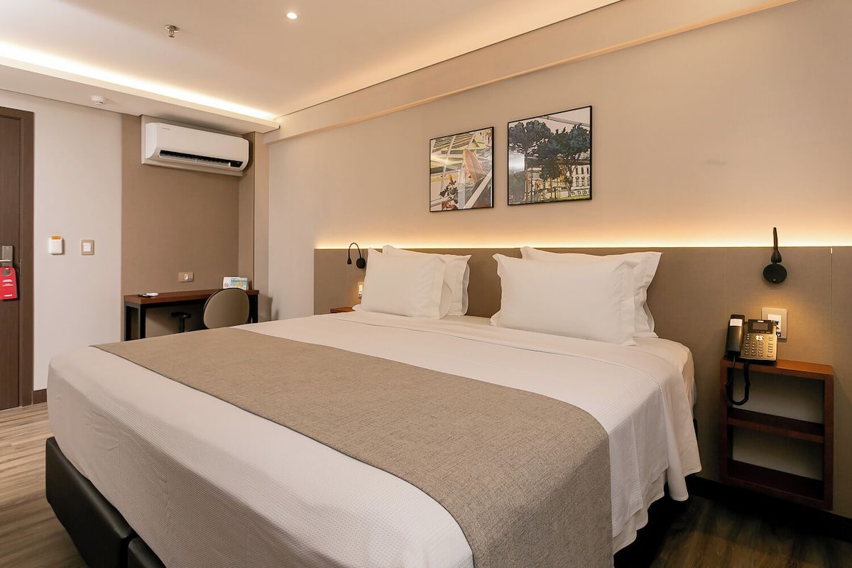 Une chambre d’hôtel avec un grand lit, un bureau, un appareil de climatisation et deux œuvres d’art accrochées au mur.