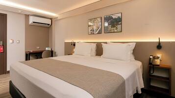 A hotel room with a large bed, a desk, an air conditioning unit, and two wall-mounted artworks.