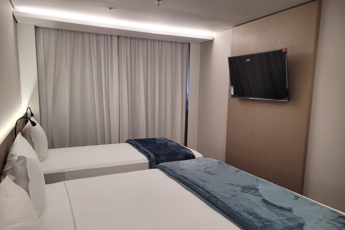 A hotel room with two beds, a television on the wall, and a window with curtains.