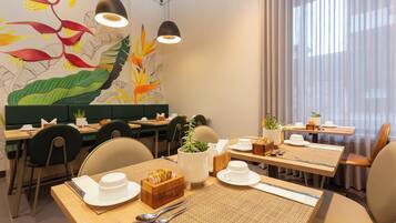 Un restaurante con un mural de aves y hojas tropicales, mesas de madera con platos y cubiertos blancos, y asientos tapizados de verde.