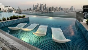 A rooftop infinity pool with lounge chairs, a city skyline view, and a calm water body.