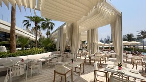 An outdoor dining area with white tables and chairs, a sandy floor, and a view of palm trees and a city skyline in the background.