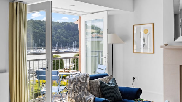 A modern living room with a blue sofa, a glass door leading to a balcony with a view of boats and greenery. - The Masts 37 Dart Marina (Dartmouth)