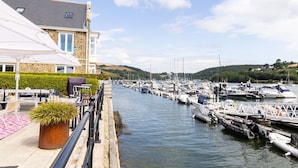 A marina with boats docked, a building with a stone facade, and a paved walkway with outdoor seating. - The Masts 37 Dart Marina (Dartmouth)