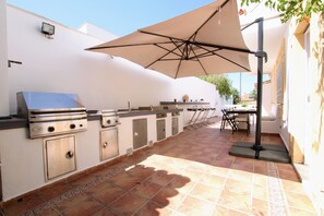 An outdoor kitchen area with a grill, bar, and chairs under a large umbrella. - Tess Villa Becerra in Alhaurin de la Torre (Alhaurin de la Torre)