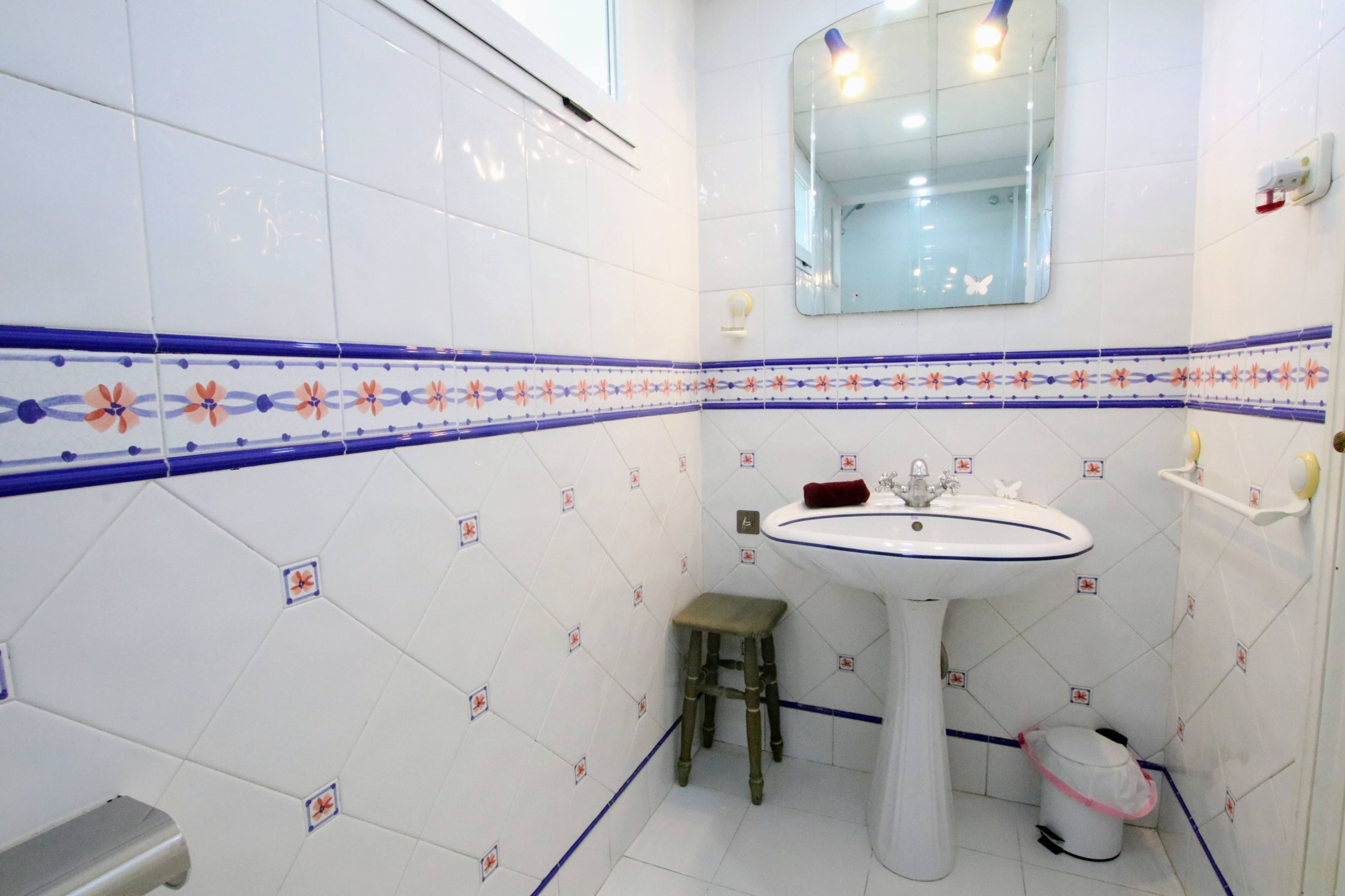 A bathroom with white tiles, a pedestal sink, a small stool, a mirror, and a shelf with toiletries.