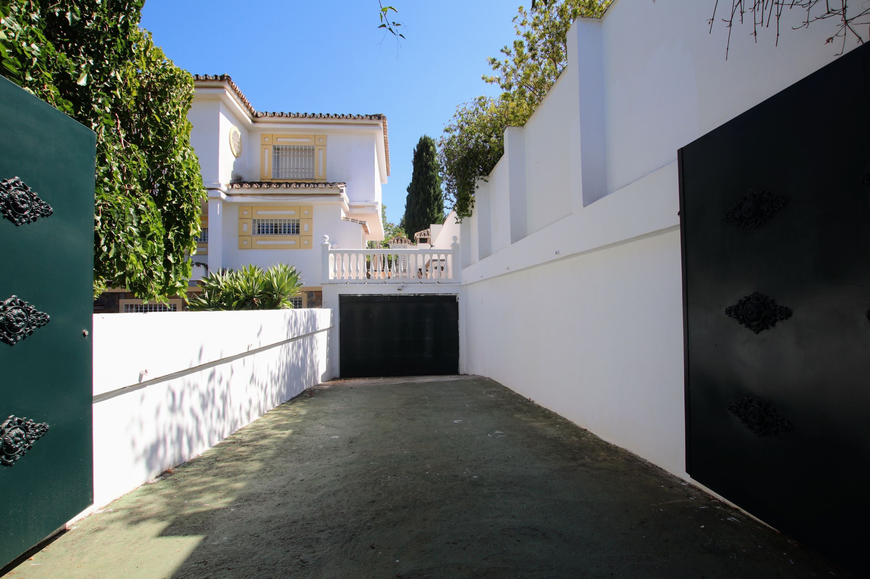 A courtyard with a white wall, a black gate, and a building with yellow trim in the background.