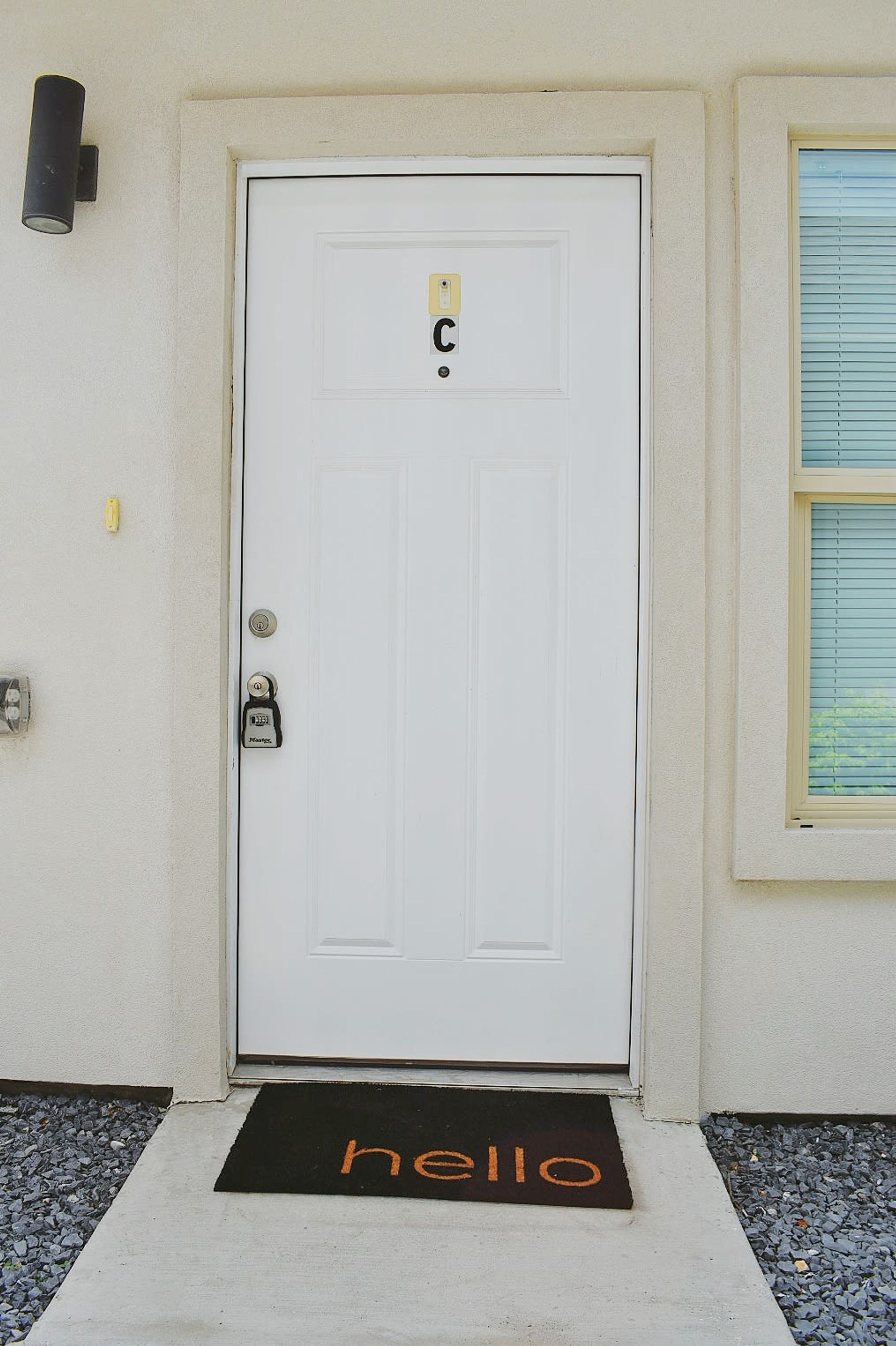 A white door with a keypad lock, a doormat with "hello" written on it, and a small window with blinds to the right.