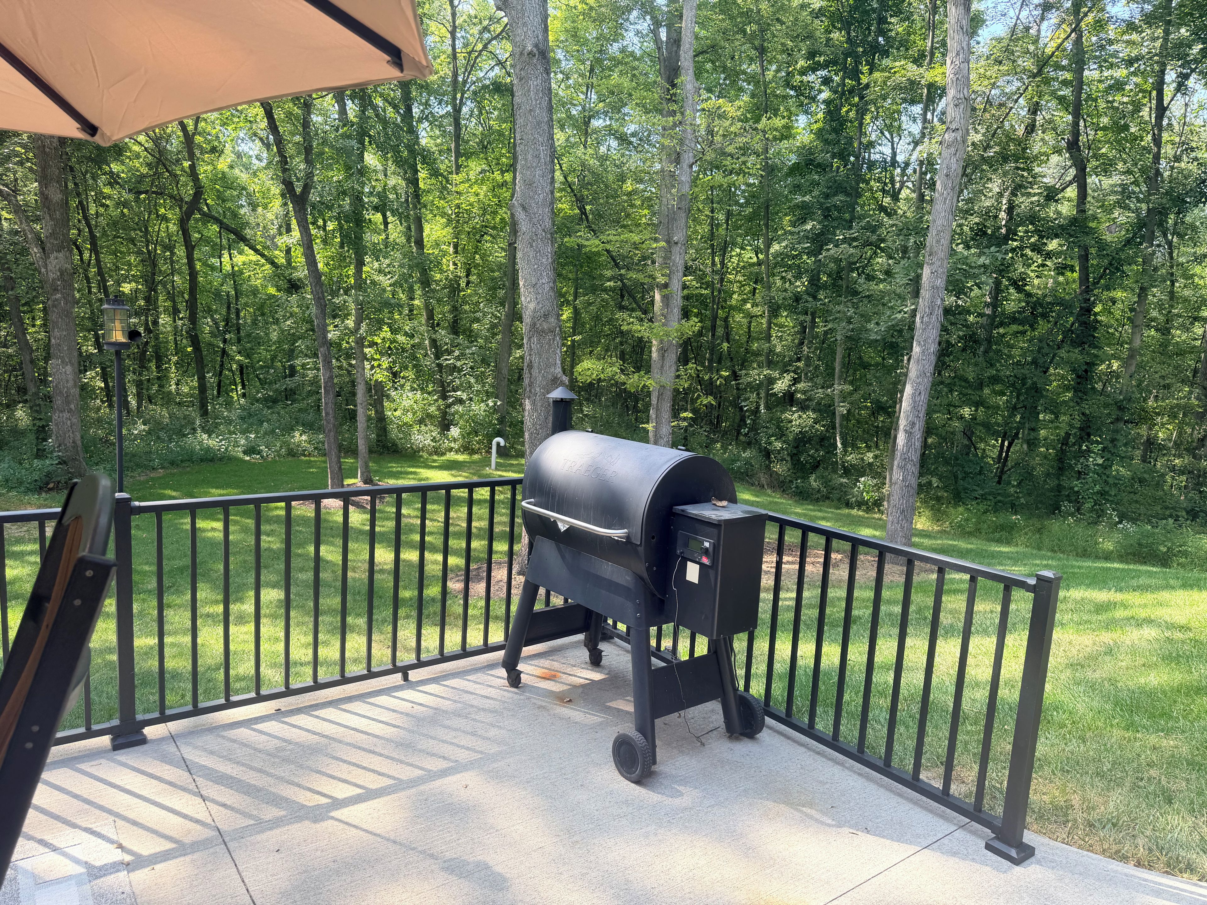 Ein Grill auf einer Terrasse mit Metallgeländer, umgeben von einer Rasenfläche und Bäumen.