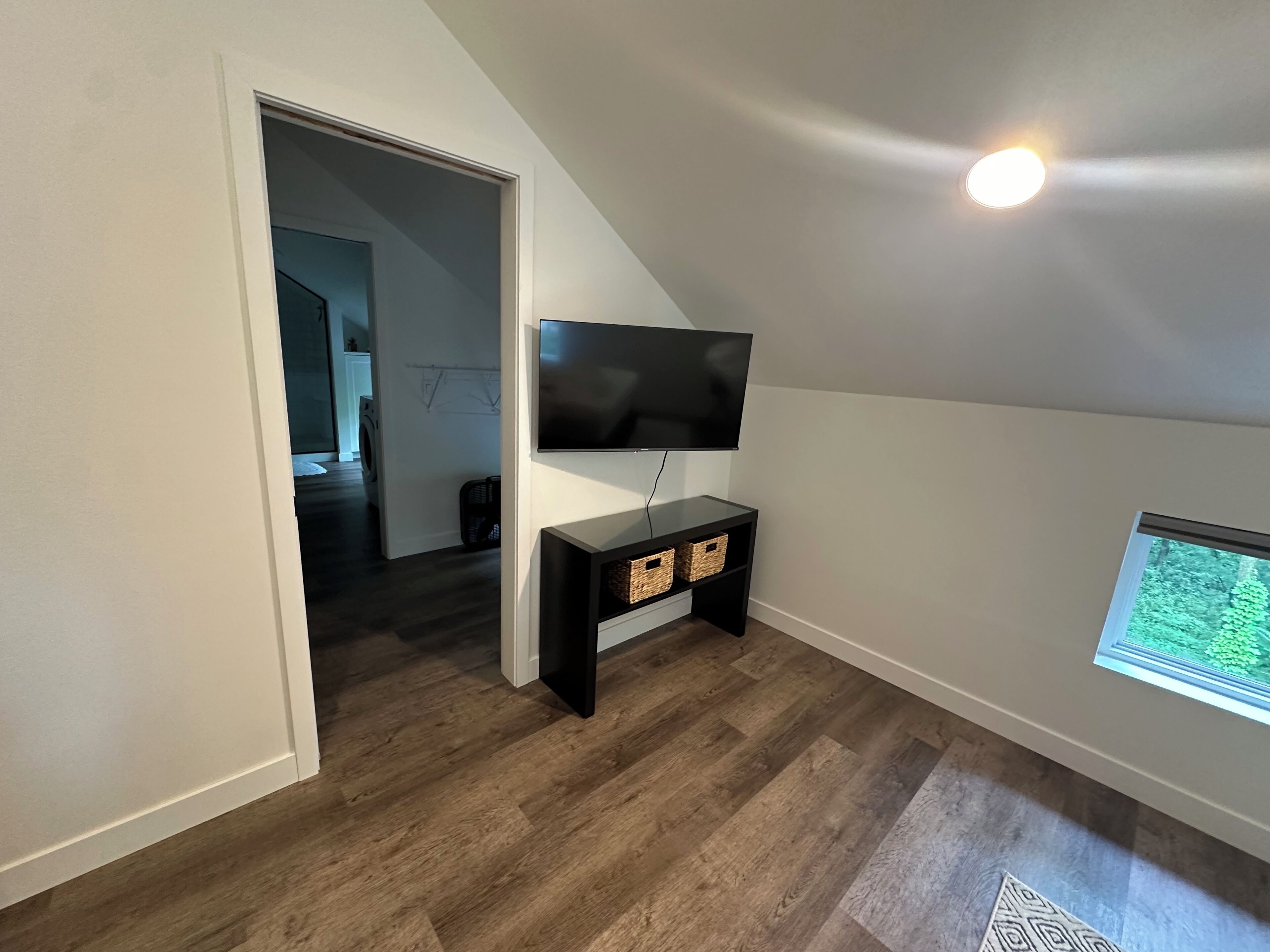 A room with a TV mounted on the wall, a wooden floor, and a window with a view of greenery.
