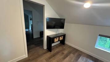 A room with a TV mounted on the wall, a wooden floor, and a window with a view of greenery.