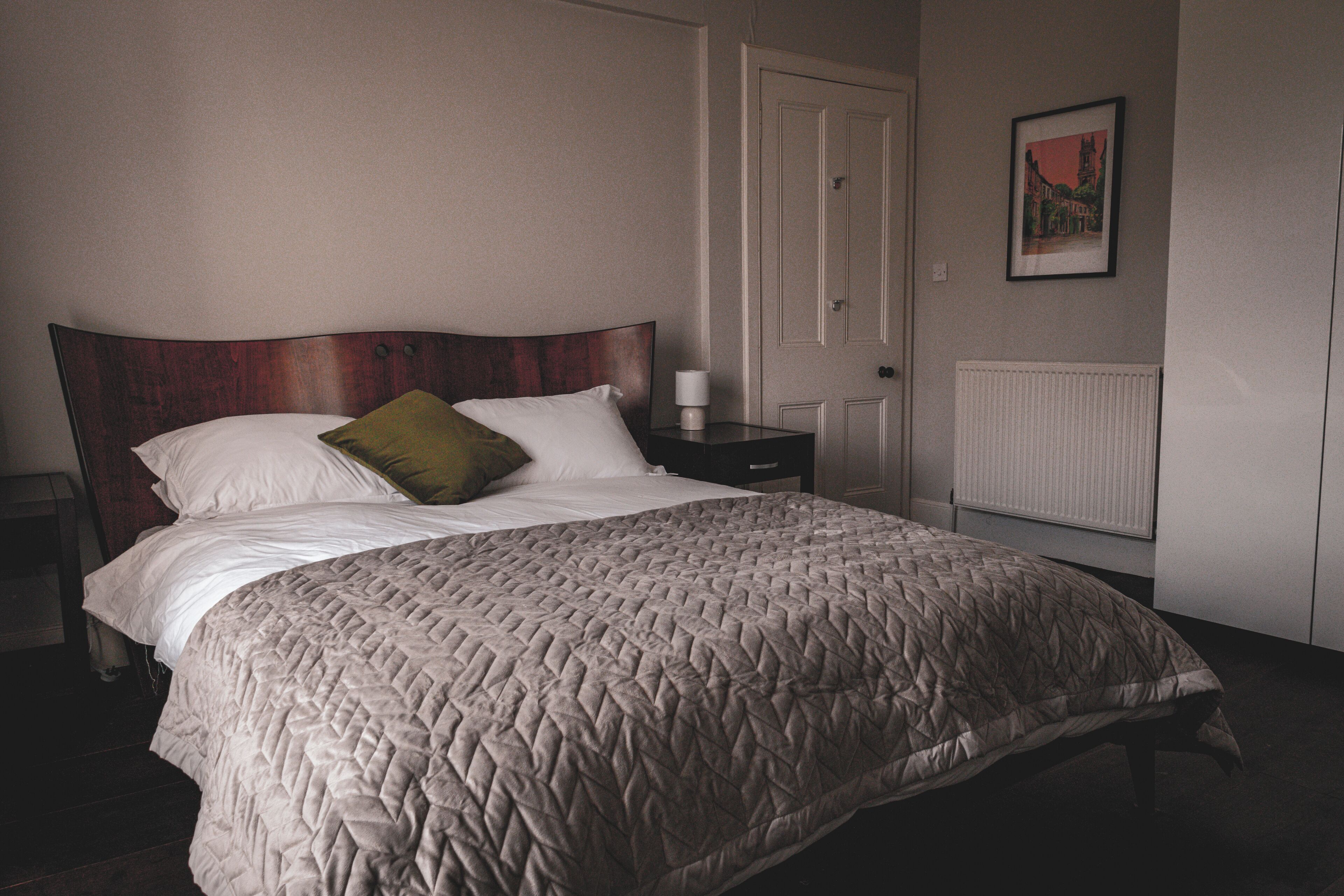 A neatly made bed with a quilted blanket, a wooden headboard, a bedside table with a lamp, and a framed picture on the wall.