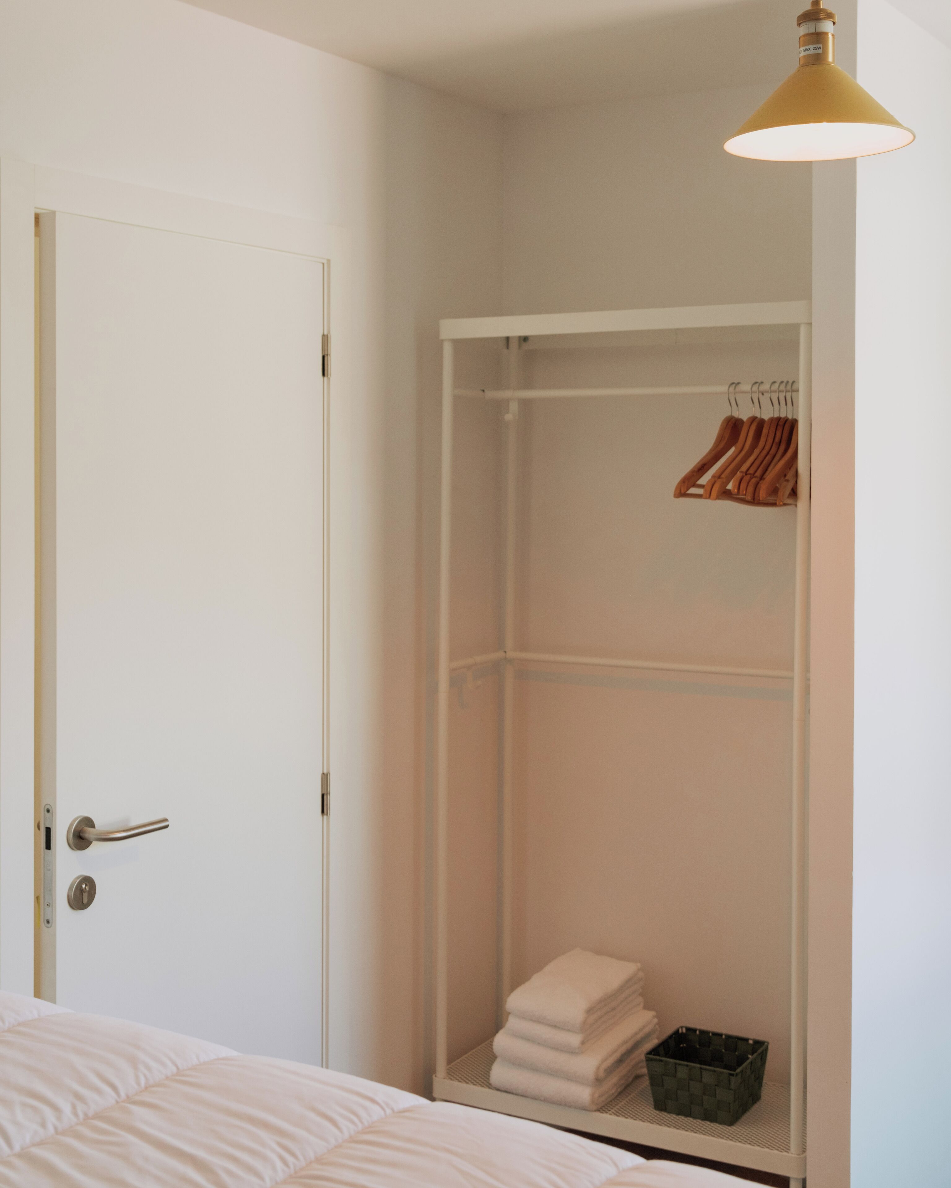 Een slaapkamer met een witte deur, een bed met witte lakens, een kledingkast met daarin hangende kleding, en een klein opbergvak.