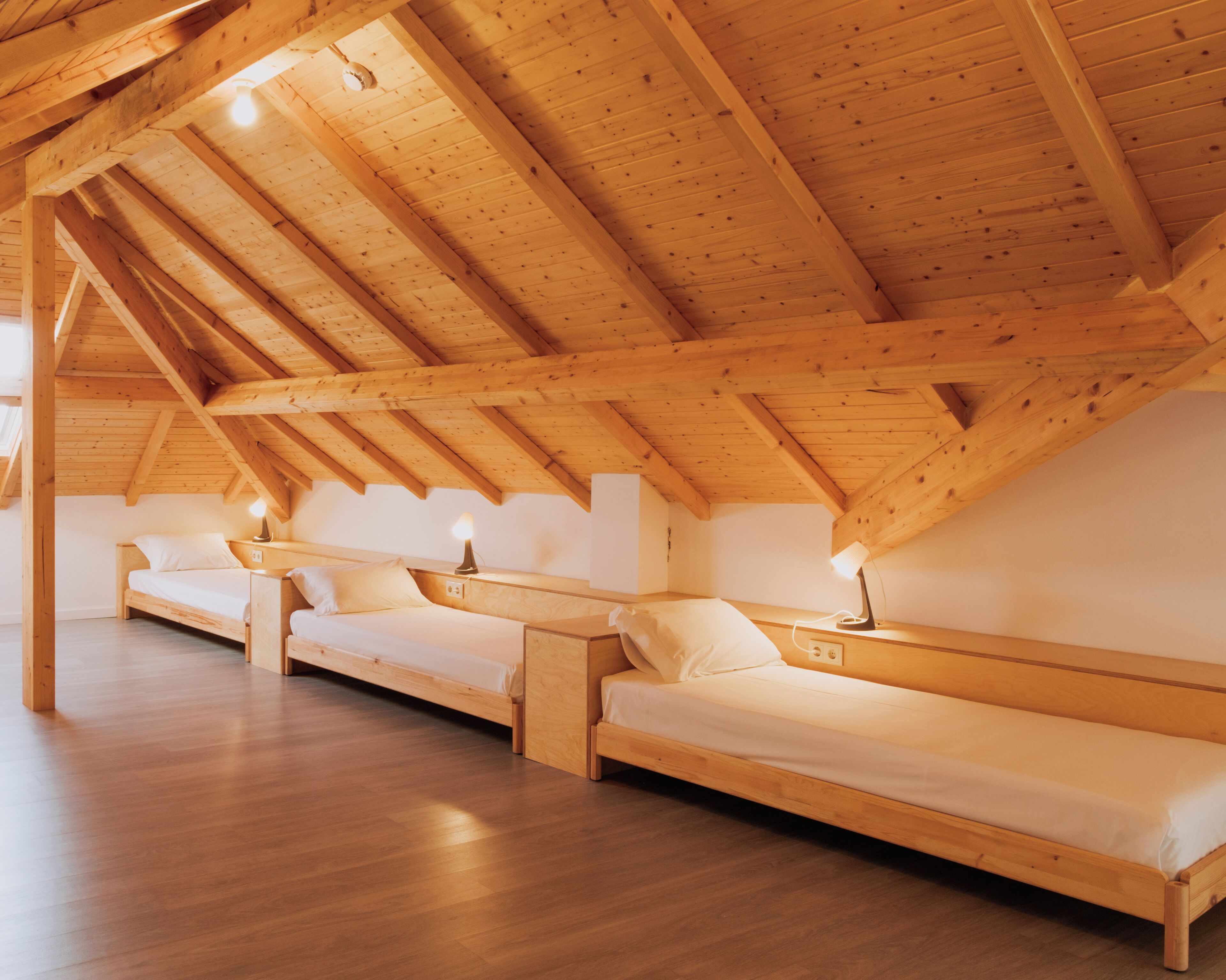 A high-ceilinged wooden room with two wooden beds and a bench, each with a bedside table and lamp.