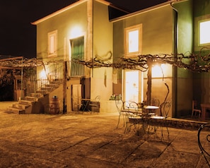 A two-story building with a staircase leading to the upper floor, outdoor seating area with metal chairs and tables, and a tree with vines. - Quinta da Quinhas (Caminha)