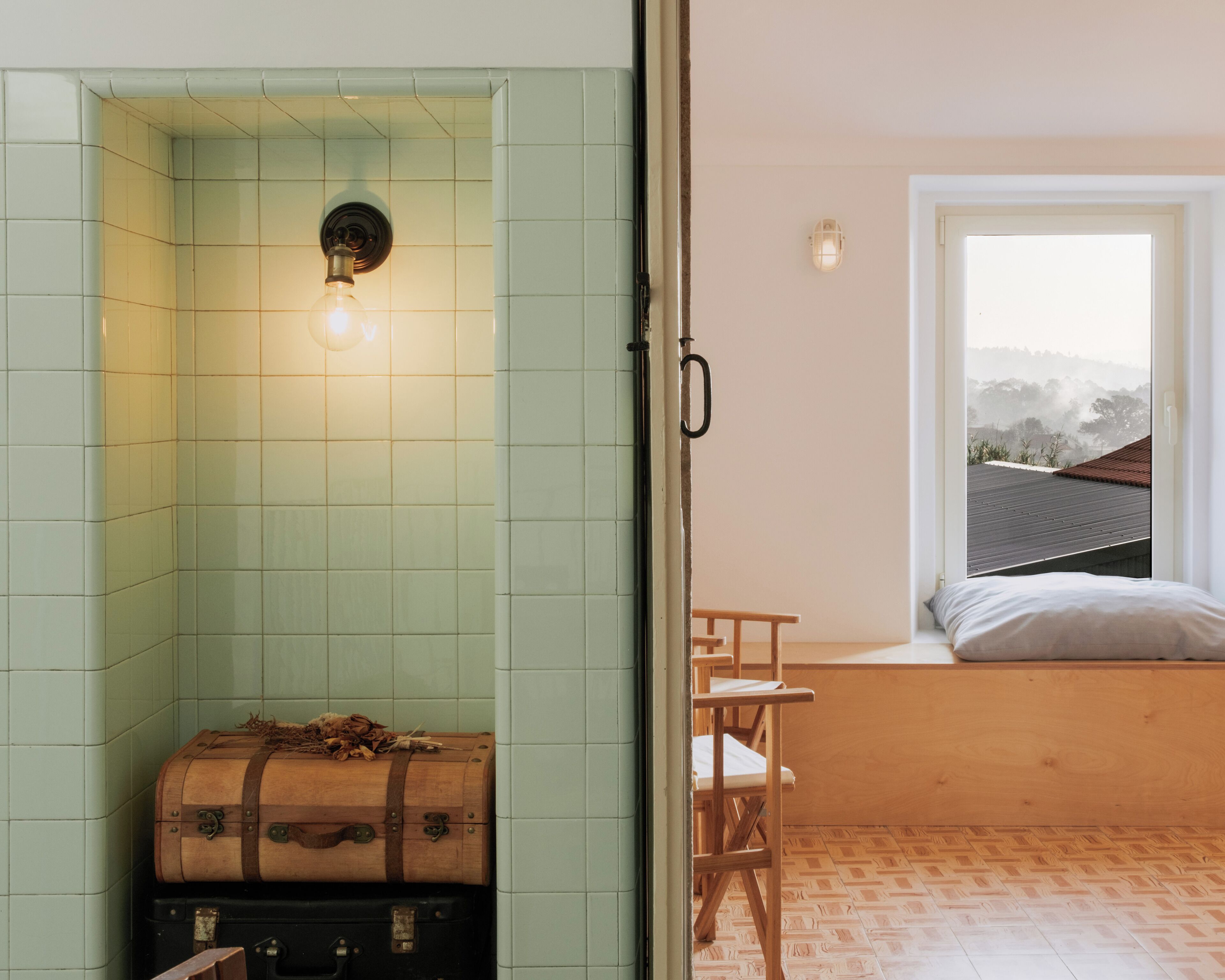 A room with a green-tiled shower, a wooden bench, a bed with a view of the outdoors, and a suitcase.