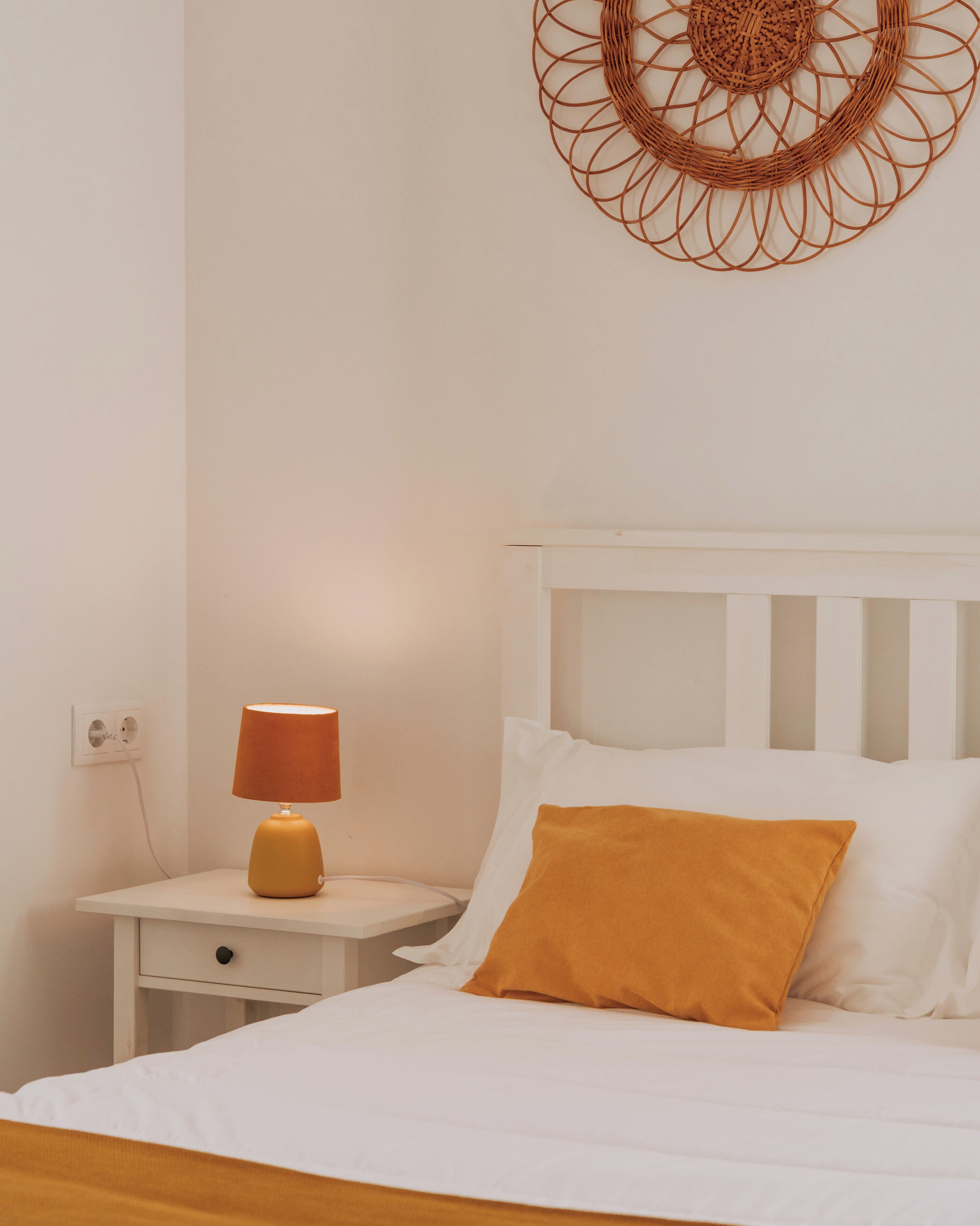 Una cama bien hecha con ropa blanca y una almohada color mostaza, una mesita de noche blanca con una lámpara y un adorno en la pared.