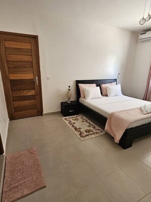 Un dormitorio con puerta de madera, cama con cabecero negro, mesita de noche con lámpara y alfombra estampada en el suelo.