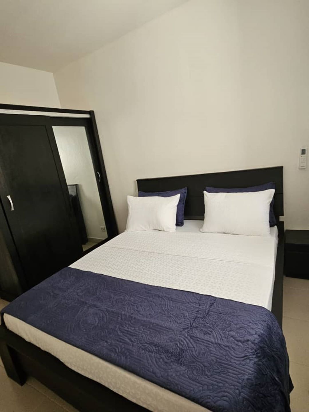 Un dormitorio con cama, cabecera oscura y pared blanca.