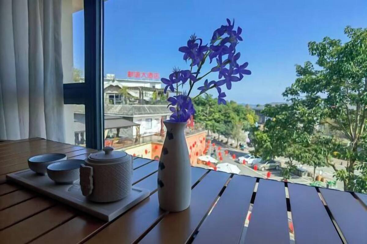 A table with a vase of purple flowers, a teapot, and two bowls on a tray, with a view of buildings and trees outside.