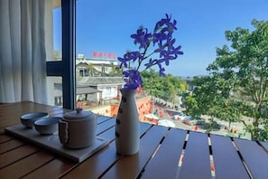 A table with a vase of purple flowers, a teapot, and two bowls on a tray, with a view of buildings and trees outside.