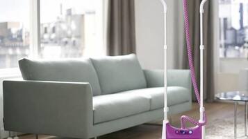 A modern living room with a light-colored sofa, a purple vacuum cleaner, and a view of a cityscape through large windows.