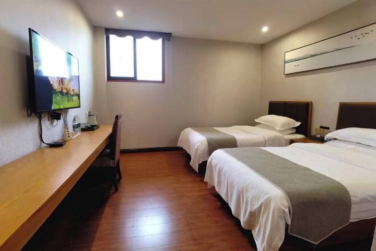 A hotel room with two beds, a TV, a window, and a wooden bench.