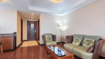 Una habitación de hotel con suelo de madera, un sofá, dos sillones, una mesa de centro con una bandeja, una lámpara y una puerta.
