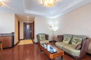 Una habitación de hotel con suelo de madera, un sofá, dos sillones, una mesa de centro con una bandeja, una lámpara y una puerta.