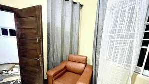 Une pièce avec un fauteuil en cuir marron, une porte en bois, des rideaux gris et une fenêtre avec un rideau blanc.