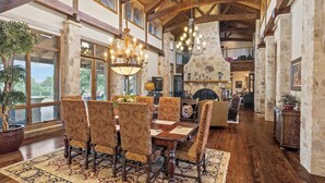 A spacious dining area with a high ceiling and wooden beams, featuring a large chandelier, patterned chairs, and a rustic stone fireplace. - Private Ranch - Pool, Ping Pong, Yoga, Weddings, Games (Bergheim)