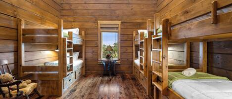 A cabin with wooden walls and floors, bunk beds, a rocking chair, and a window with a view of greenery.