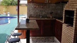 A kitchen with a stone backsplash, a built-in oven, and a poolside bar with stools. - Casa 6 pessoas com Piscina (Sao Pedro da Aldeia)