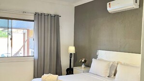 A hotel room with a bed, a ceiling fan, an air conditioner, a window with curtains, and a nightstand with a lamp and a vase of flowers. - Casa 6 pessoas com Piscina (Sao Pedro da Aldeia)