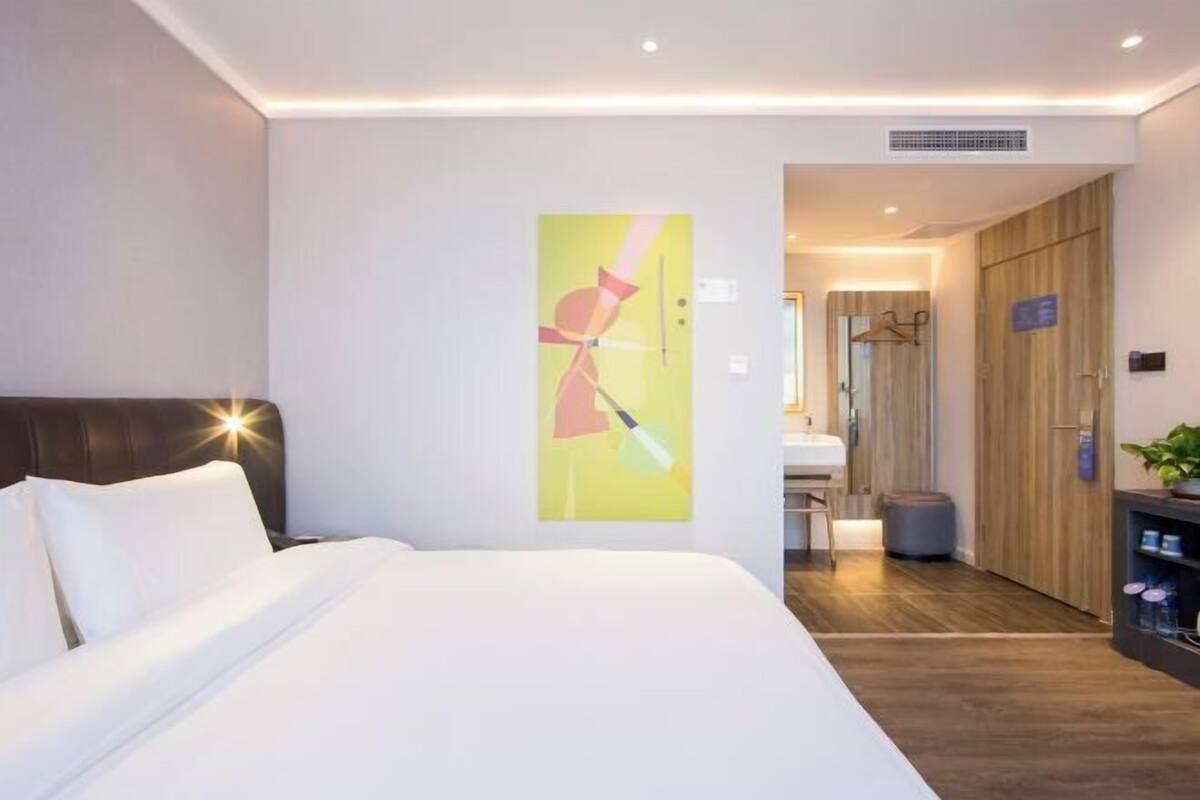A hotel room with a bed, a painting on the wall, a wooden wardrobe, and a small table with a plant.